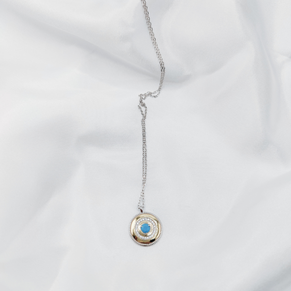 Este elegante collar presenta un colgante redondo adornado con brillantes circonitas que rodean una deslumbrante piedra azul en el centro.