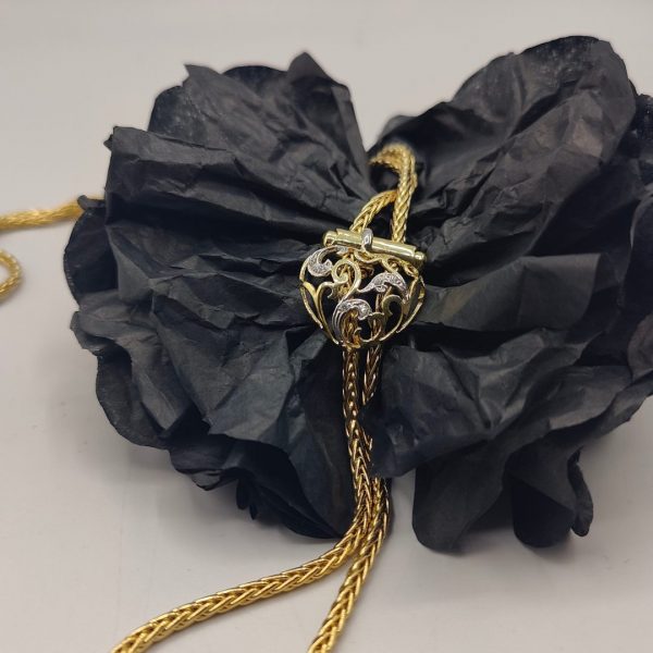 cadena estilo corbatín con eslabones tipo serpiente, cierre corazón con circonitas y el final de la cadena tiene dos borlas una de ellas lleva de circonitas