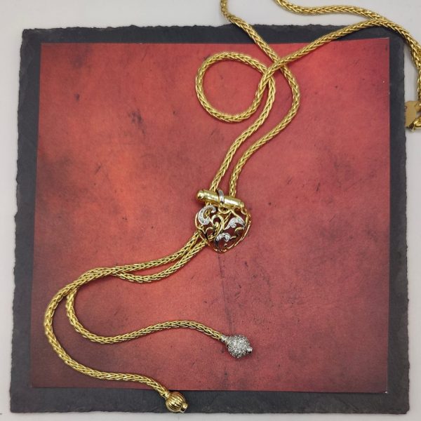 cadena estilo corbatín con eslabones tipo serpiente, cierre corazón con circonitas y el final de la cadena tiene dos borlas una de ellas lleva de circonitas