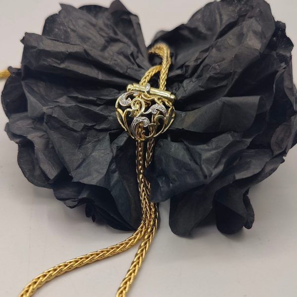 cadena estilo corbatín con eslabones tipo serpiente, cierre corazón con circonitas y el final de la cadena tiene dos borlas una de ellas lleva de circonitas